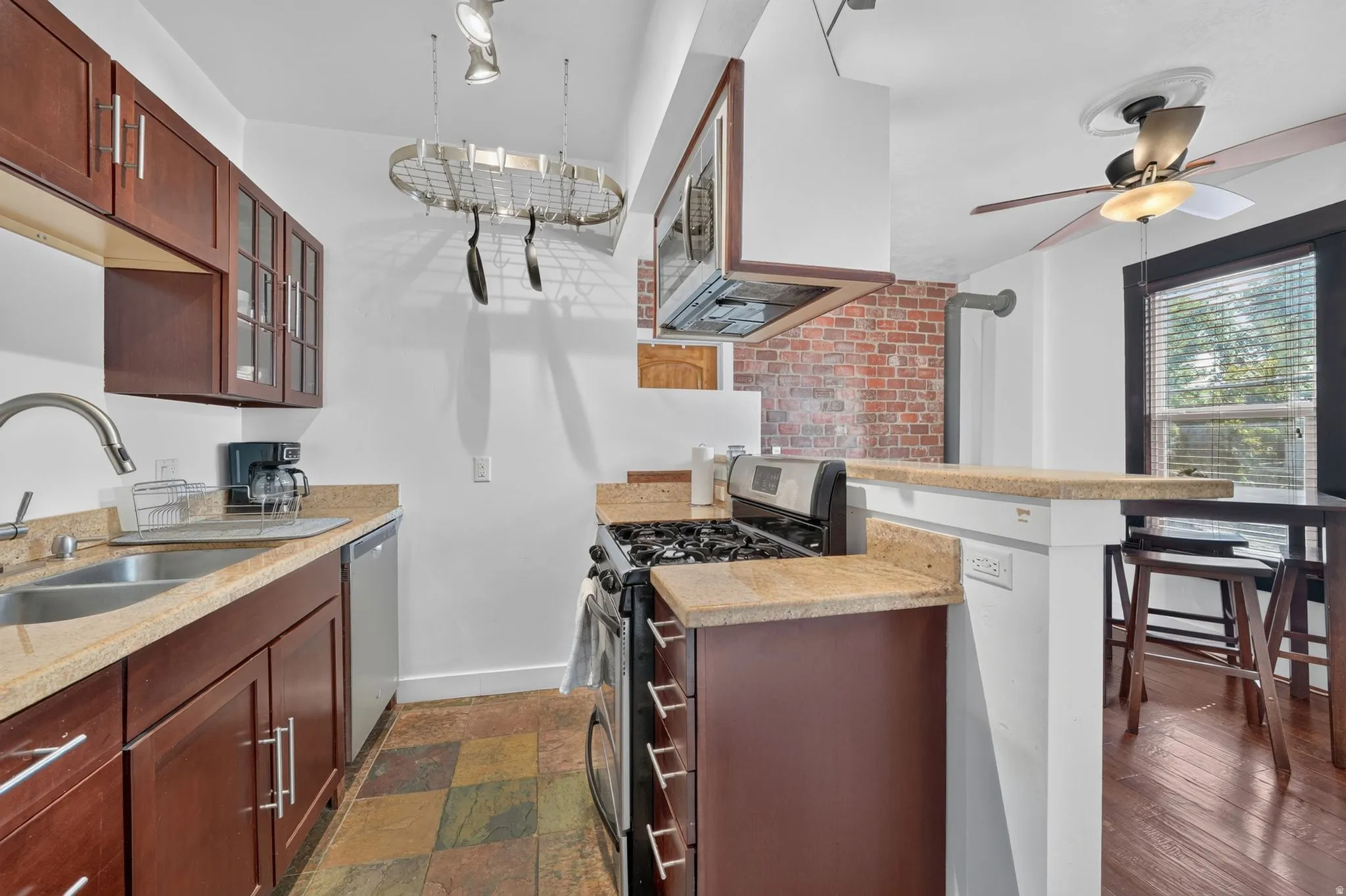 Kitchen featuring rich wood cabinetry with glass-front accents, light-toned countertops, a stainless steel double basin sink with a gooseneck faucet, a pot rack, and multi-toned slate-look flooring