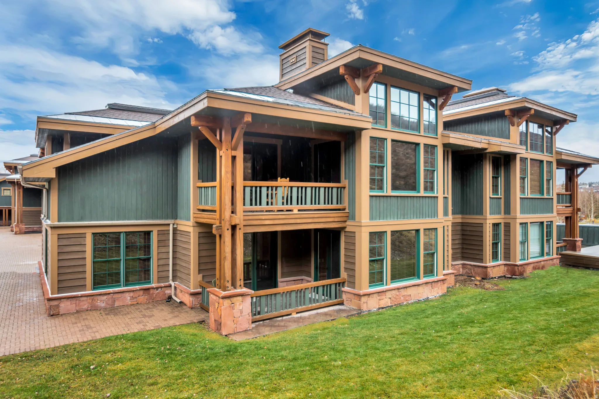 Multi-story building featuring green and brown siding, exposed timber framework, multiple balconies, a brick-paved patio, and a sloped lawn