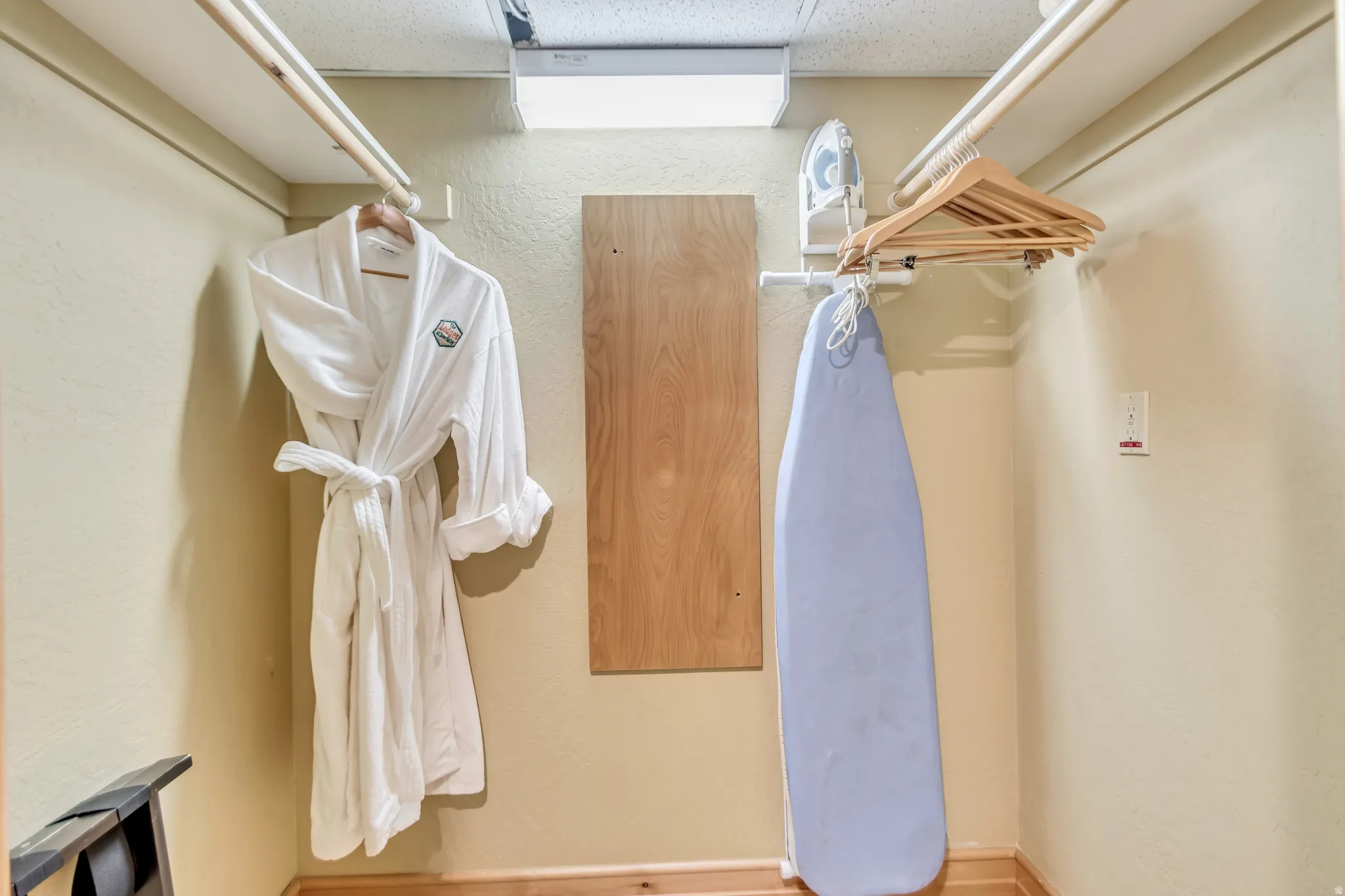 Compact utility closet featuring a wall-mounted ironing board, integrated hanging rods, and a light-toned wood-finish floor