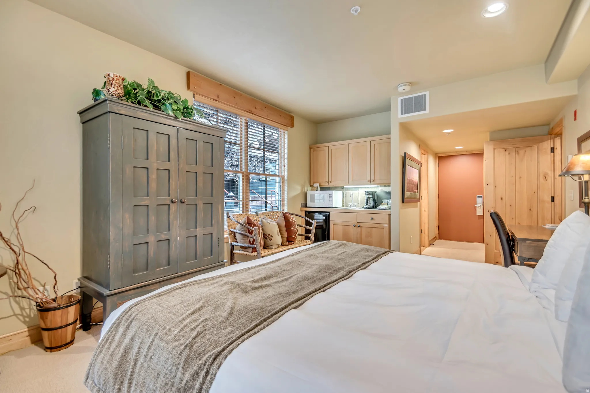 Compact living space featuring a built-in kitchenette with wood-finish cabinetry, a window with horizontal blinds, recessed lighting, and a desk area with wood-finish paneling