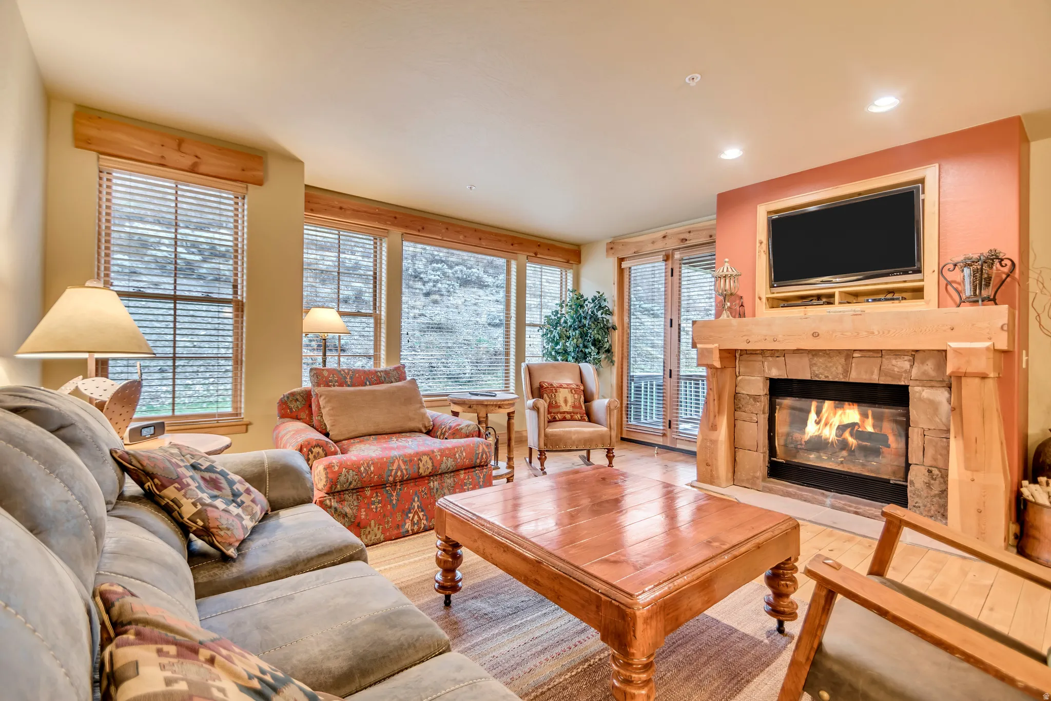 Living area featuring wood-finish flooring, a stone-surround fireplace with a wood mantle, and multiple windows providing natural light