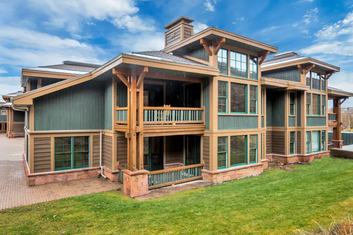 Multi-story building featuring green and brown siding, exposed timber framework, multiple balconies, a brick-paved patio, and a sloped lawn