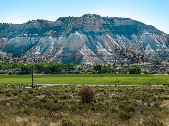 Expansive mountain landscape featuring layered rock formations, varied natural foliage, and a broad green field with irrigation systems