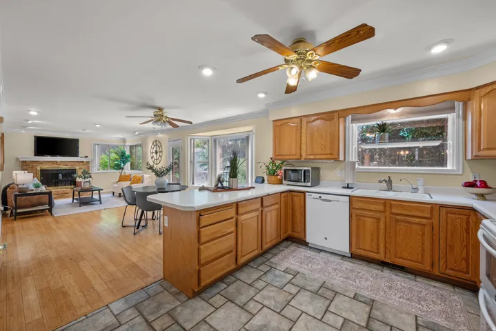 Open-concept living area featuring a kitchen with wood cabinetry and tile flooring, a living room with wood-finish flooring, a brick-surround fireplace, and multiple windows