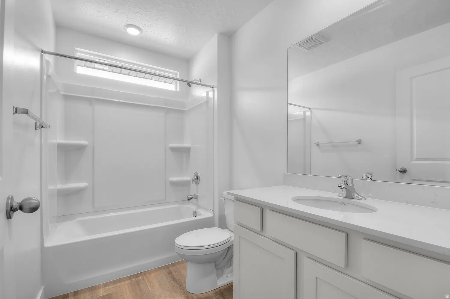 Bathroom featuring a white integrated tub and shower unit with built-in shelving, a large vanity mirror, a single-sink vanity with a light-colored countertop, white cabinetry, and wood-finish flooring