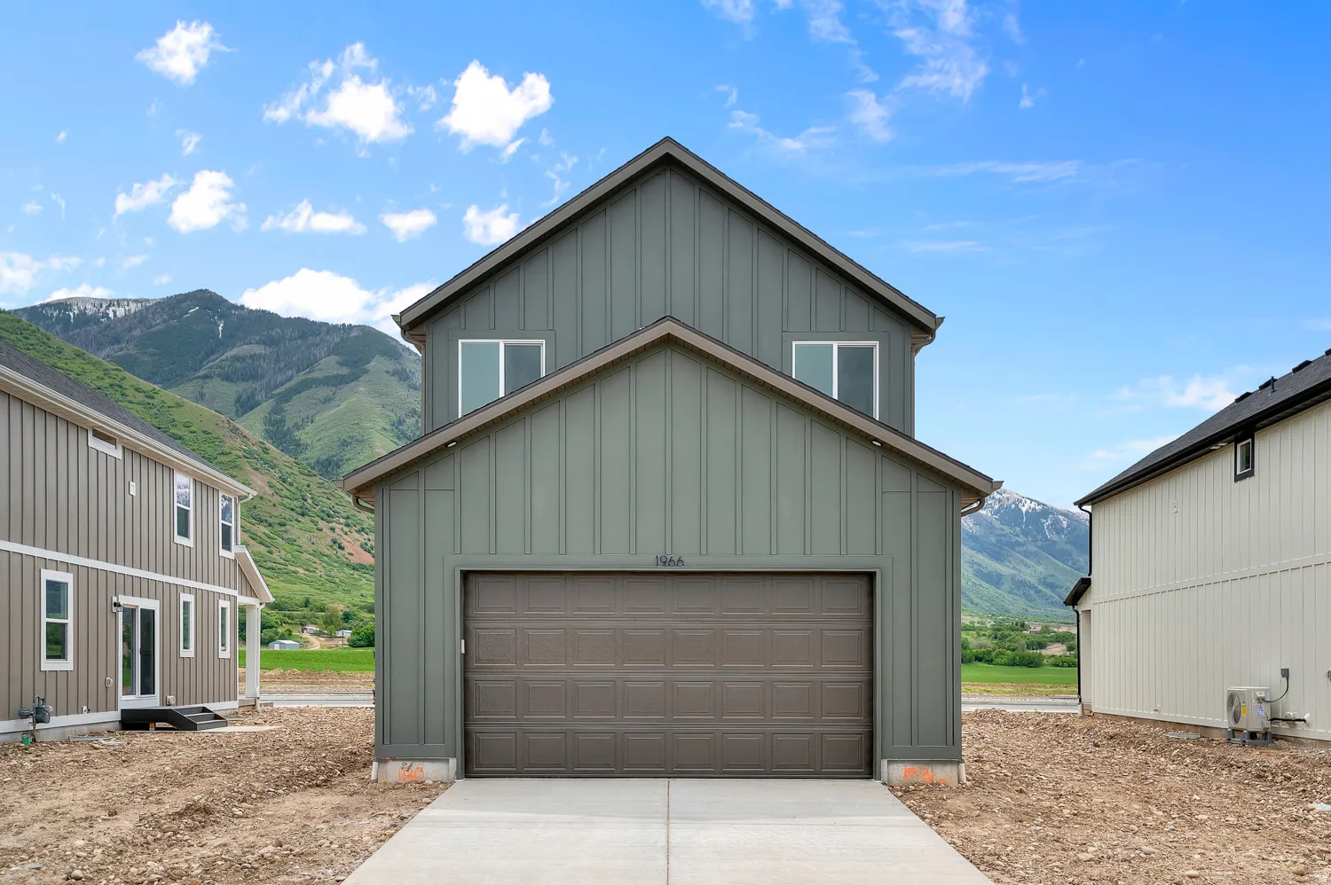 Modern farmhouse-style exterior featuring vertical siding in a muted green hue, a two-car garage with a panel door, and prominent mountain scenery in the background