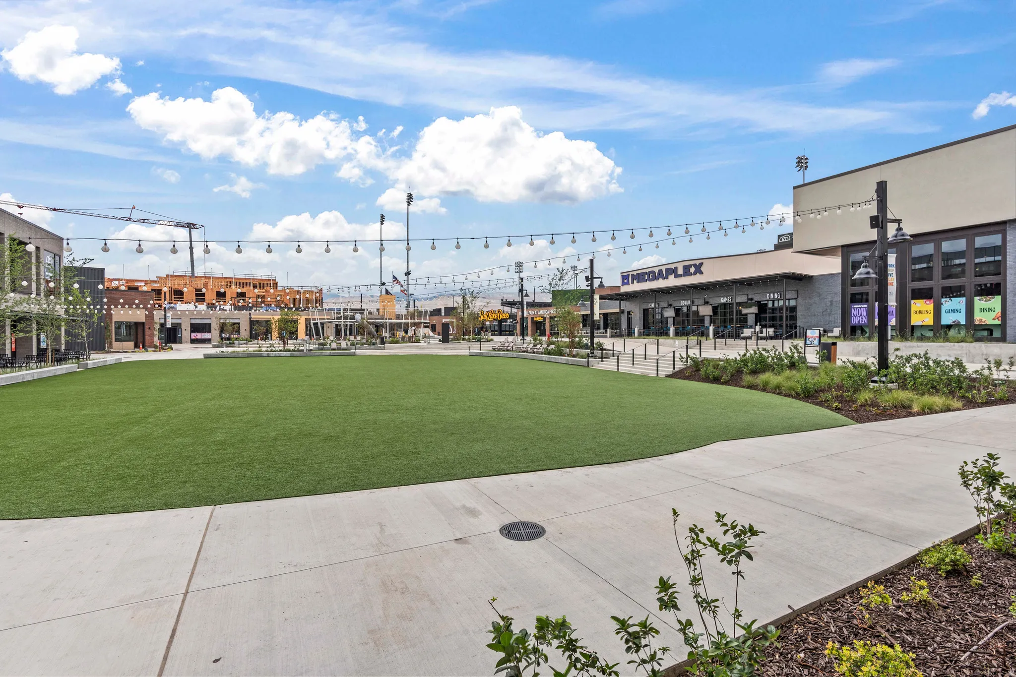 Expansive green space featuring a manicured lawn, surrounding paved walkways, string lighting overhead, and various commercial buildings