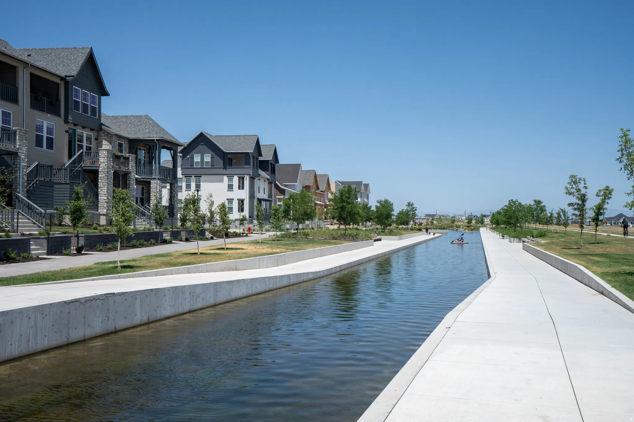 Contemporary multi-level residences featuring balconies and stone-accented exteriors bordering a waterway with concrete pathways and landscaped green spaces