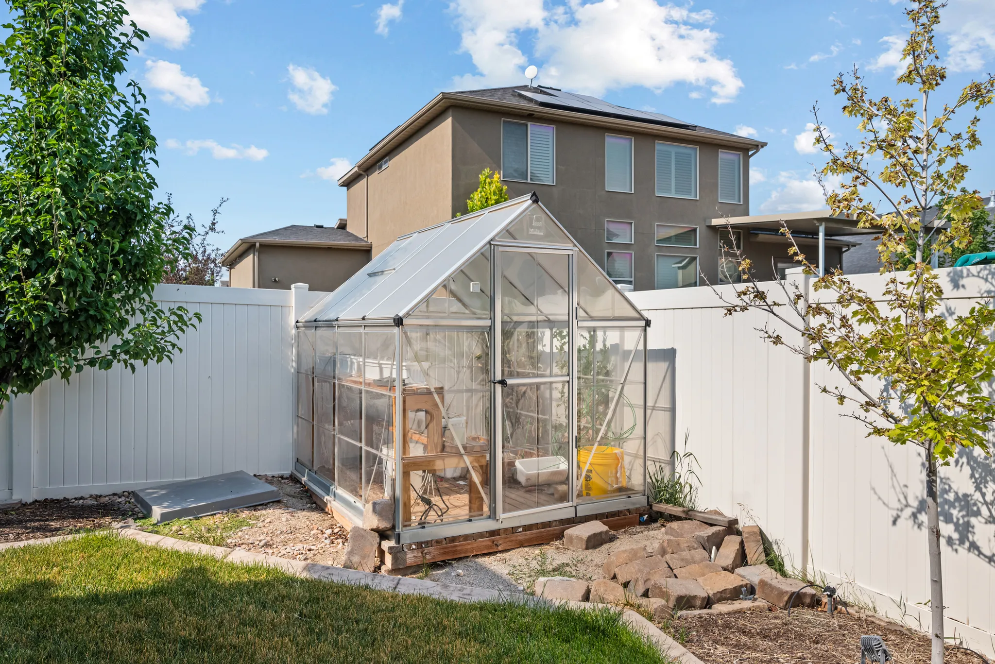 Rear yard featuring a clear-paneled greenhouse with a pitched roof, a white vinyl privacy fence, and mature trees