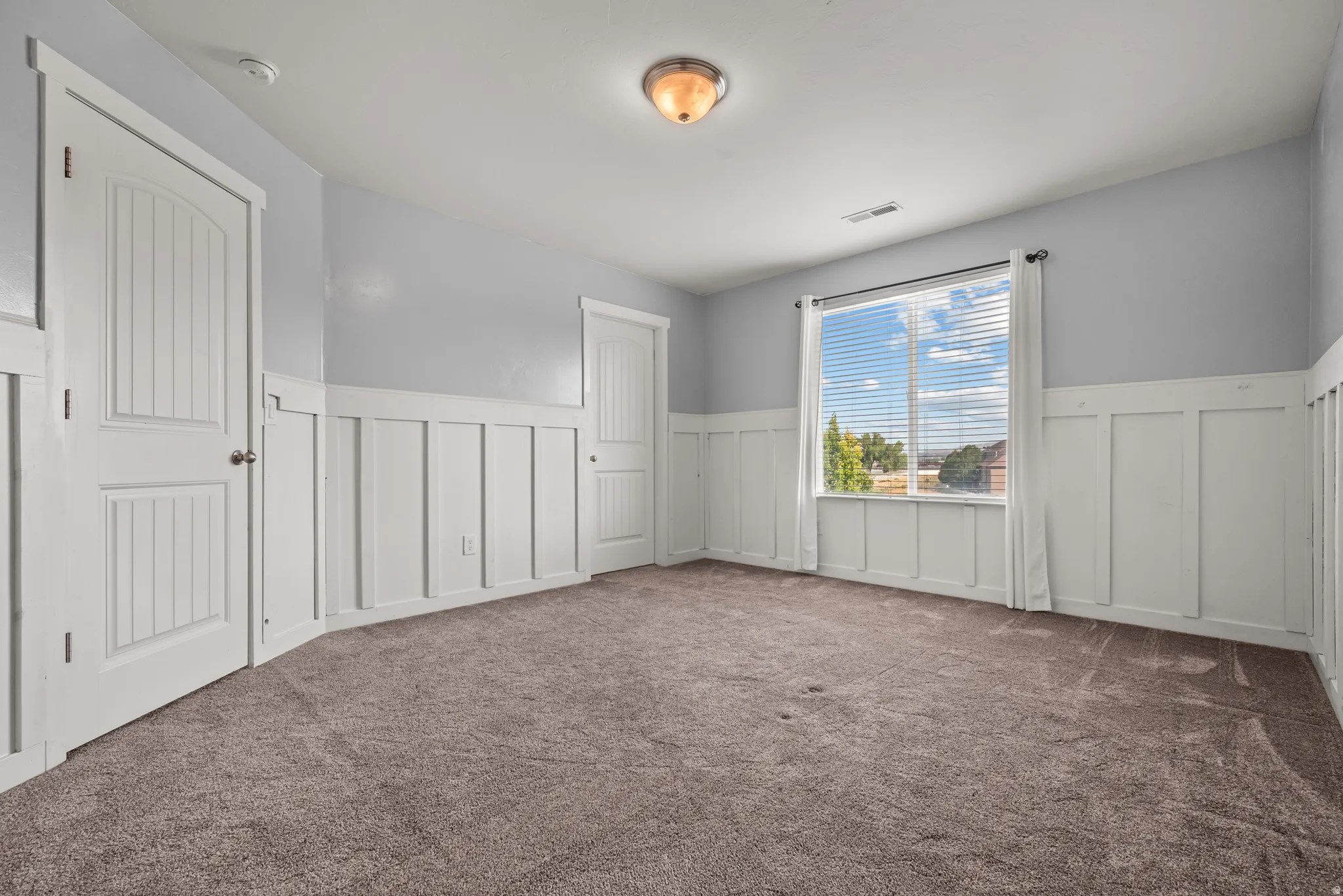 Carpeted room featuring a large window with blinds, white wainscoting, two paneled doors, and a ceiling-mounted light fixture