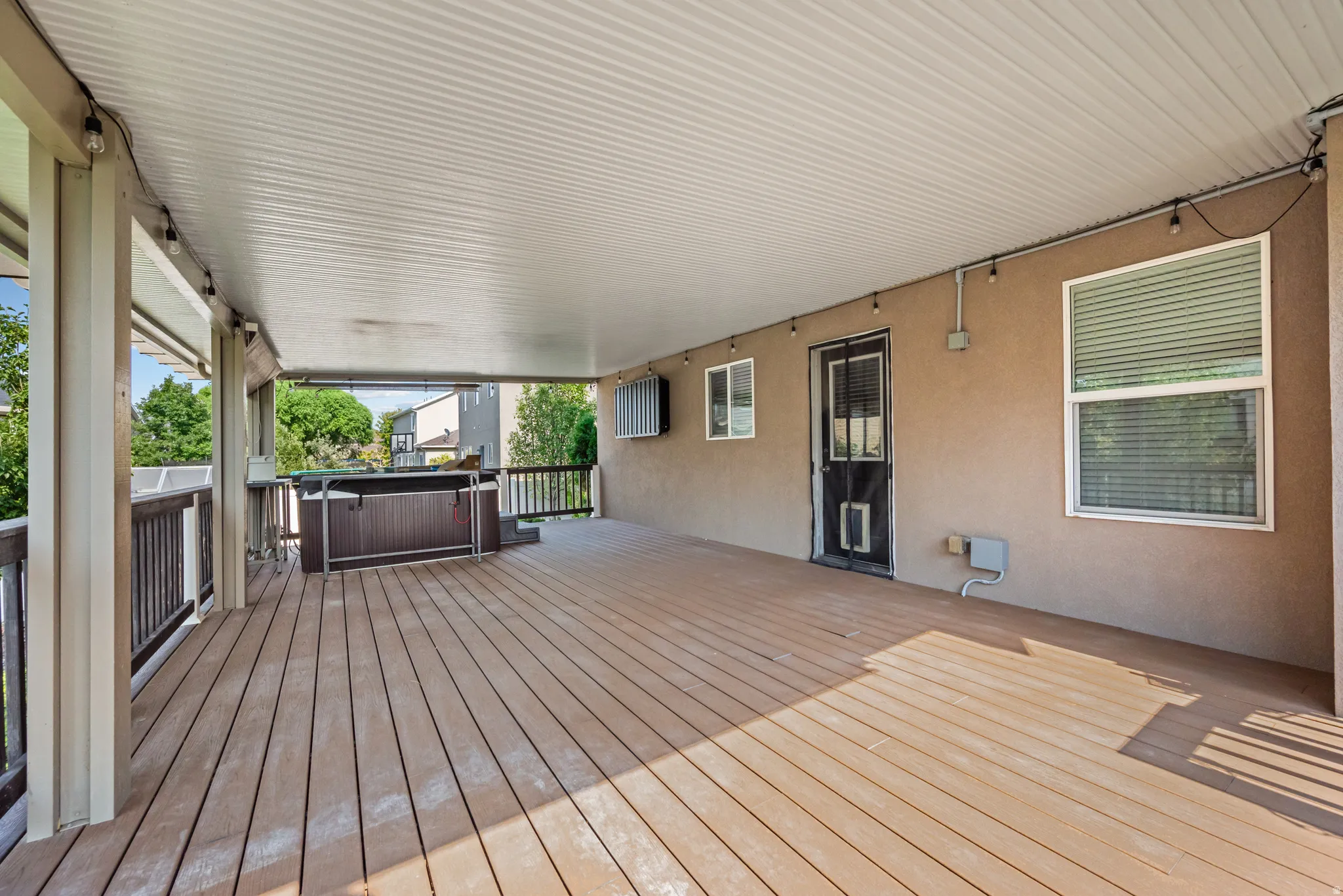 Expansive covered deck featuring wood-finish flooring, a built-in hot tub, and a stucco exterior wall with an exterior door and multiple windows