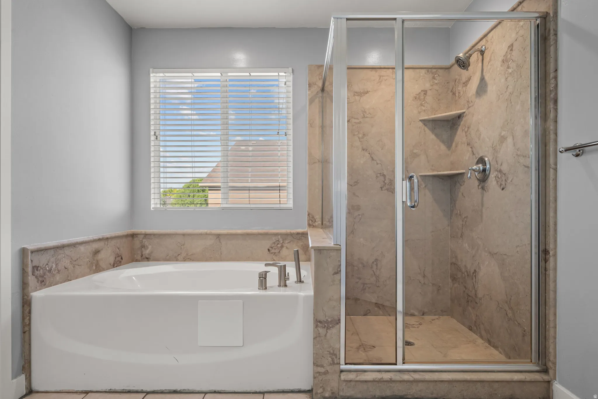 Spacious bathroom featuring a built-in soaking tub with deck-mounted fixtures, a separate glass-enclosed shower with stone-look tile surround and built-in shelving, and a window with blinds