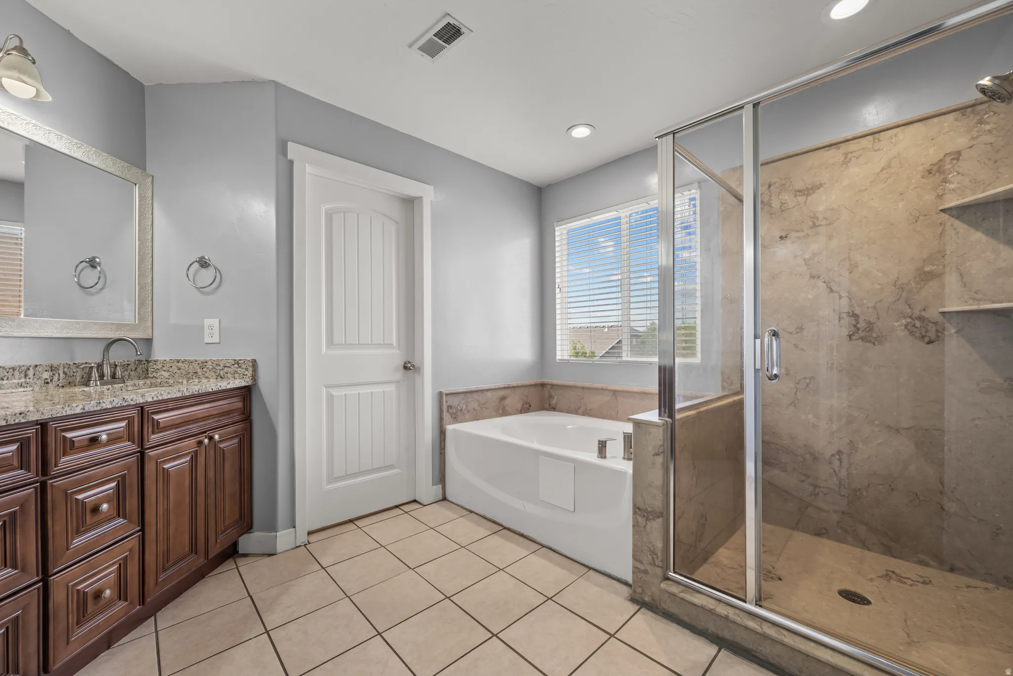 Bathroom featuring a spacious walk-in shower with stone-look tile, a separate built-in soaking tub, a double vanity with a stone countertop, and tile flooring