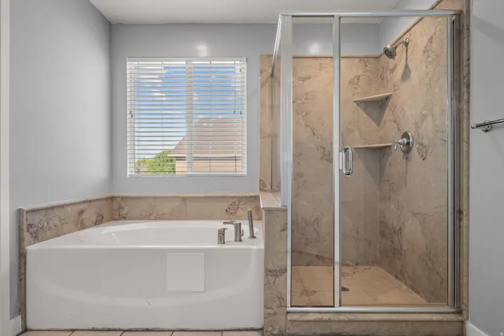 Spacious bathroom featuring a built-in soaking tub with deck-mounted fixtures, a separate glass-enclosed shower with stone-look tile surround and built-in shelving, and a window with blinds