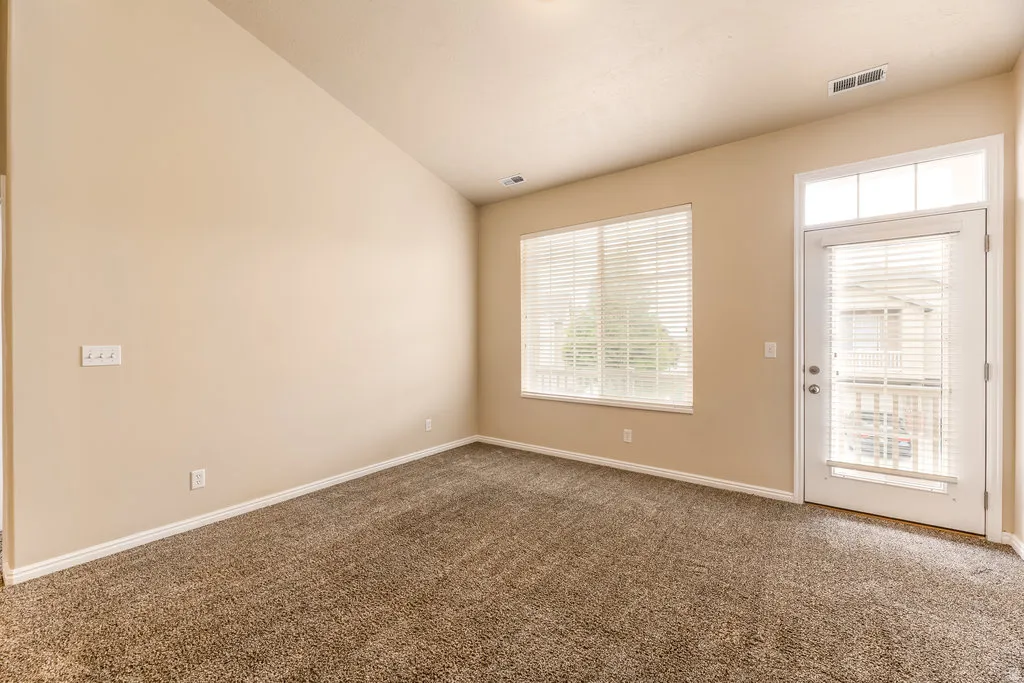 Carpeted interior space with a window featuring horizontal blinds, a white door with a transom window above, and light beige wall paint