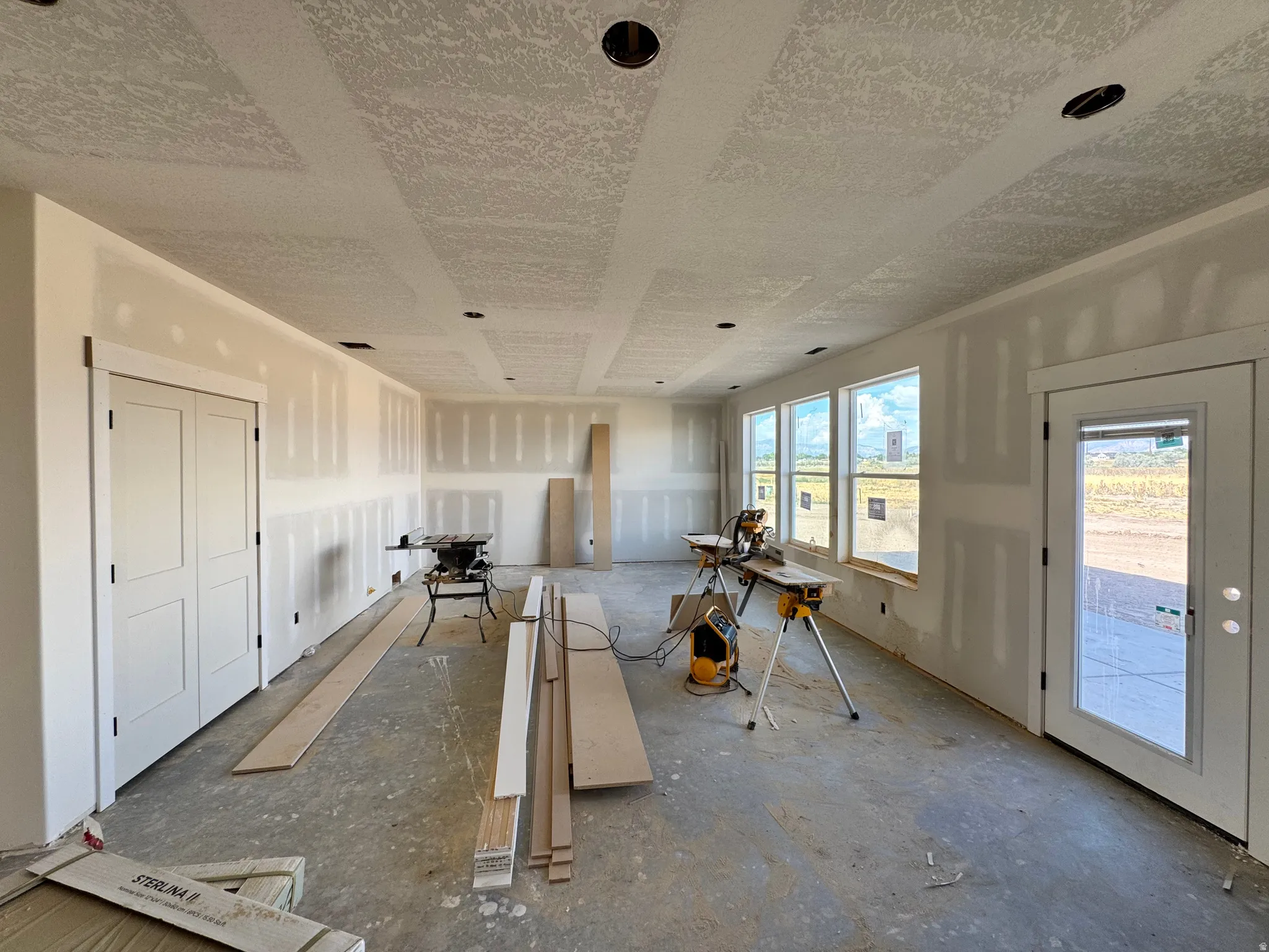 Expansive interior with unfinished drywall, multiple windows, recessed ceiling light fixtures, and a glass-paneled exterior door