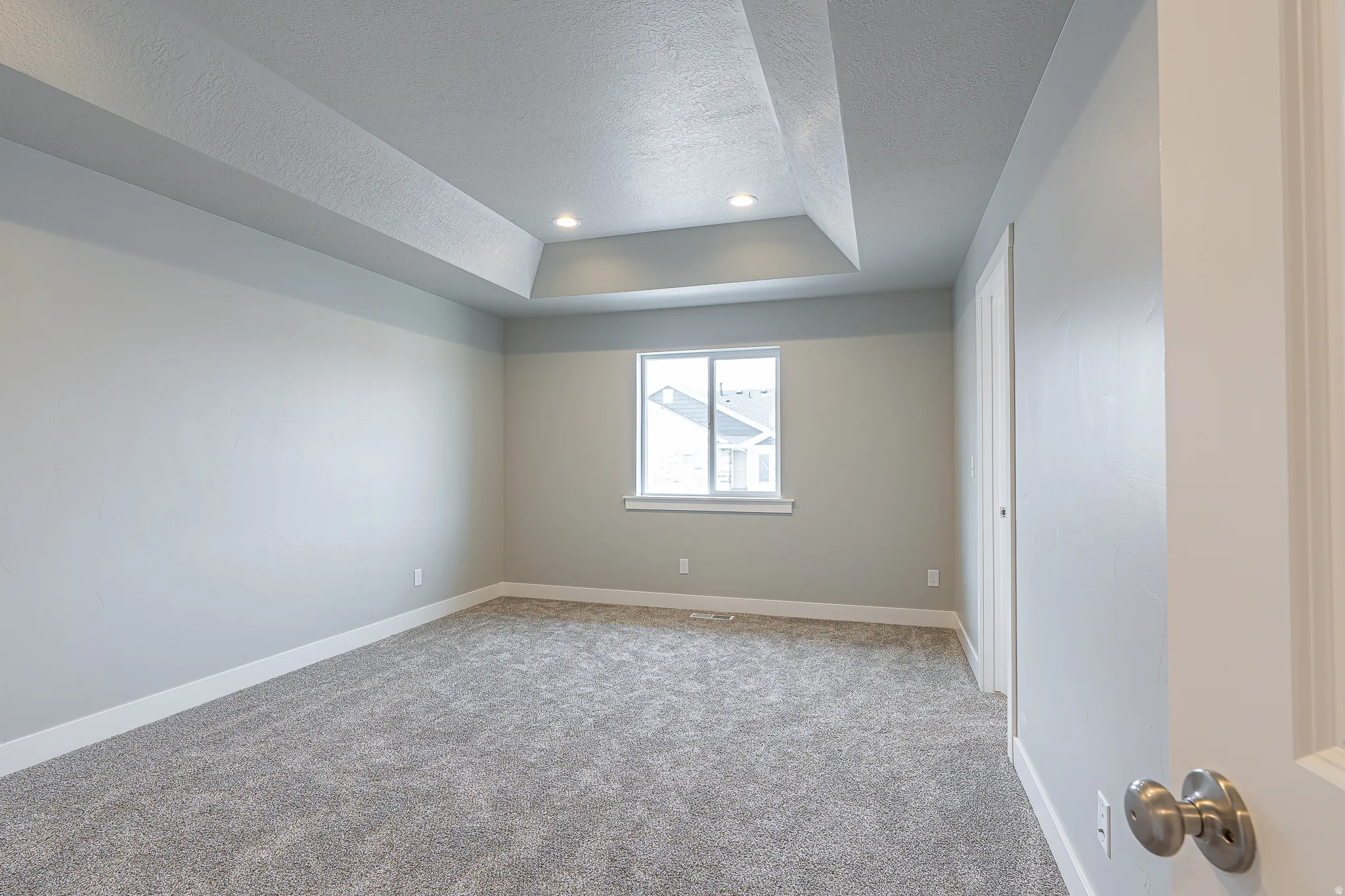 Spacious room featuring a tray ceiling with recessed lighting, light gray walls, a window with white trim, and gray carpet flooring