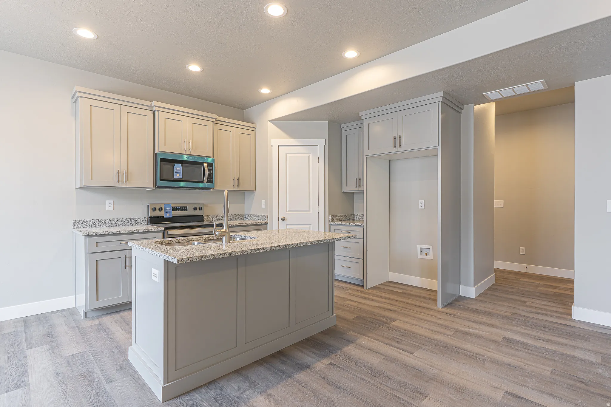 Kitchen featuring an island with a stainless steel sink, light grey cabinetry, granite countertops, and wood-finish flooring