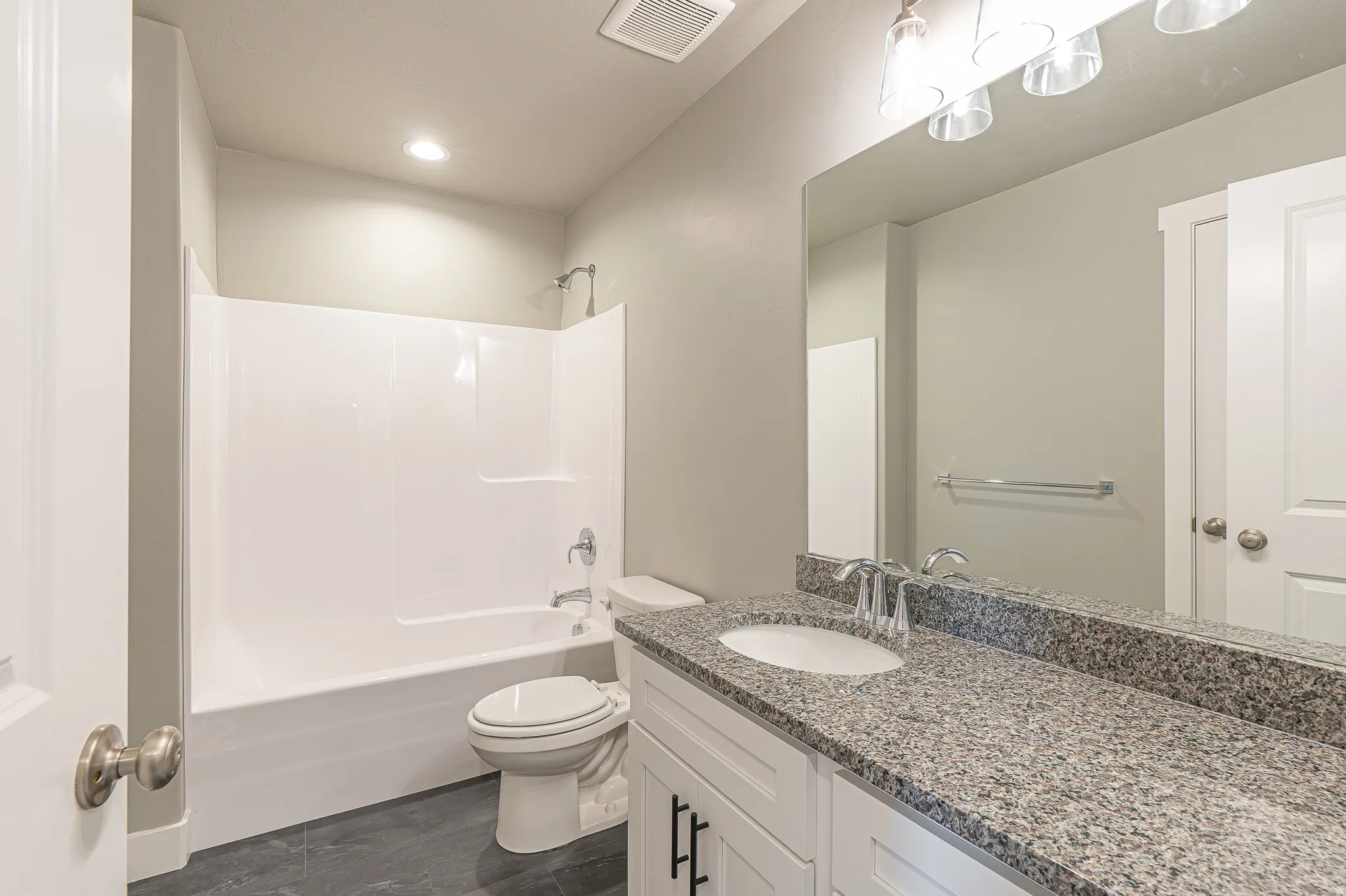 Bathroom featuring a white tub/shower combination, dark tile flooring, a single vanity with a granite-finish countertop, undermount sink, and a large mirror