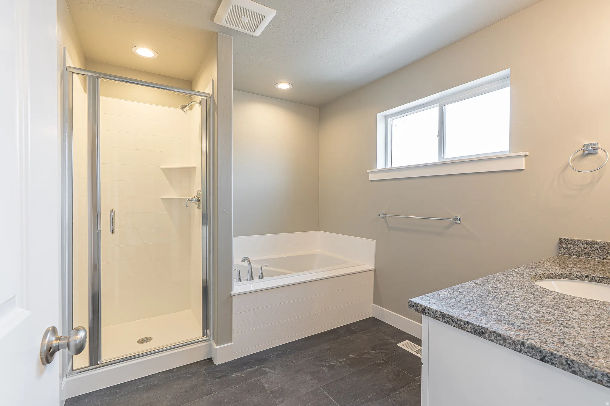 Full bathroom featuring a walk-in shower with clear glass enclosure, a built-in soaking tub, a single vanity with stone-finish countertop, and dark tile flooring