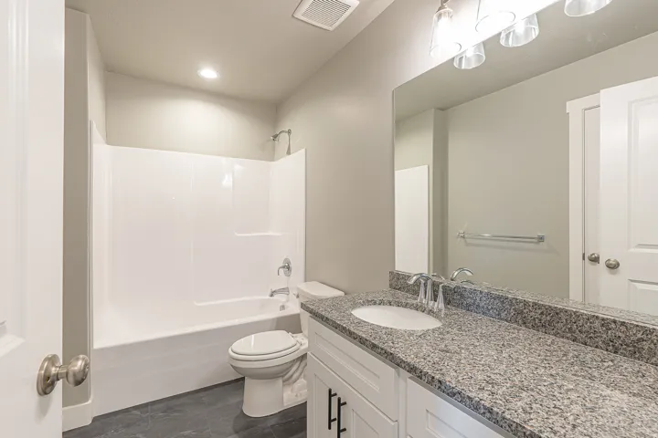 Bathroom featuring a white tub/shower combination, dark tile flooring, a single vanity with a granite-finish countertop, undermount sink, and a large mirror
