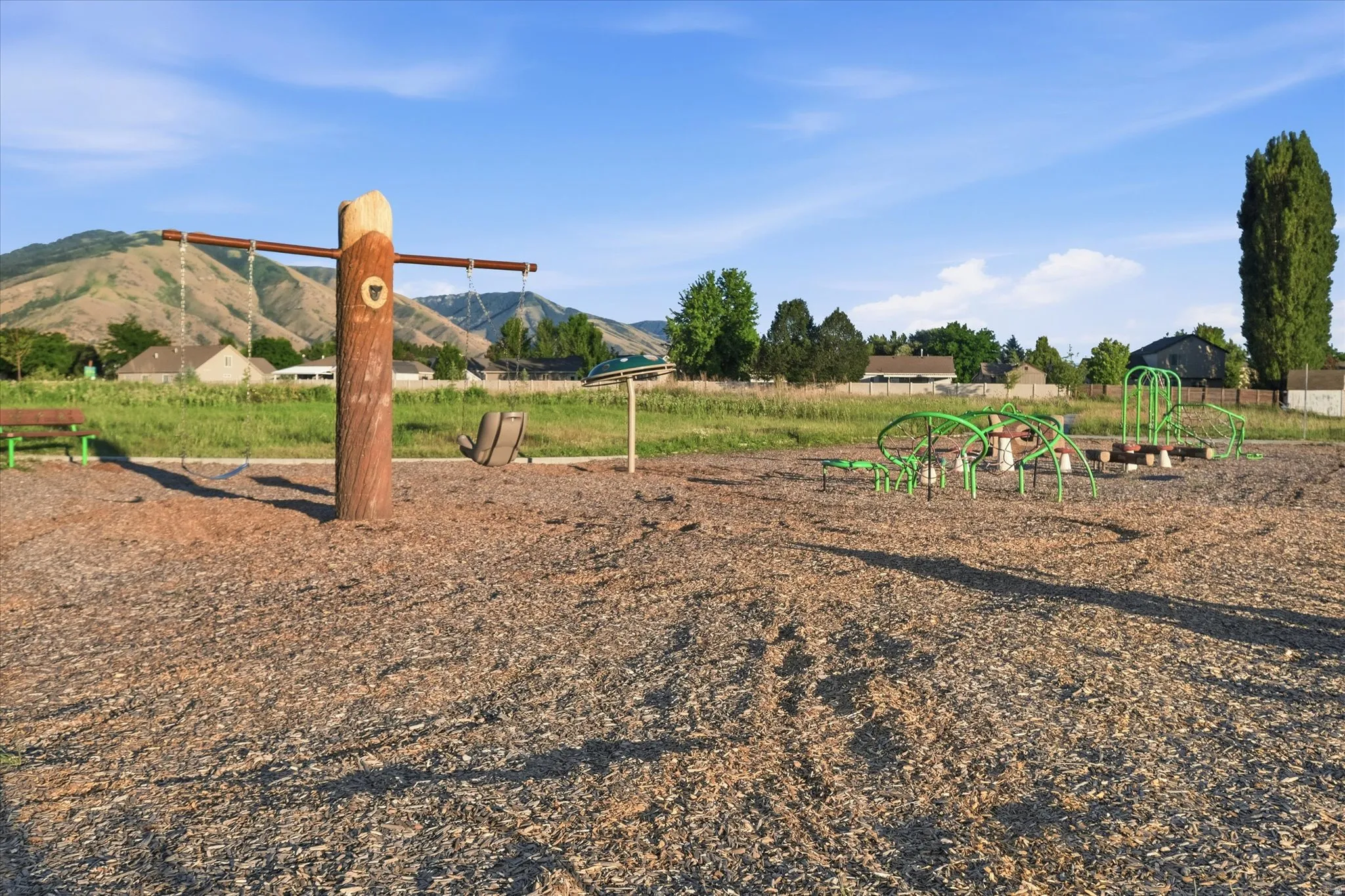 Outdoor playground featuring a swing set with a wood-look support post, green-framed climbing structures, and a wood chip ground cover
