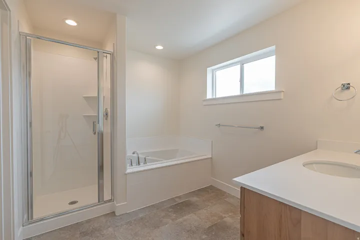Spacious bathroom featuring a glass-enclosed shower, built-in soaking tub, wood-finish vanity with an undermount sink, and light-toned flooring