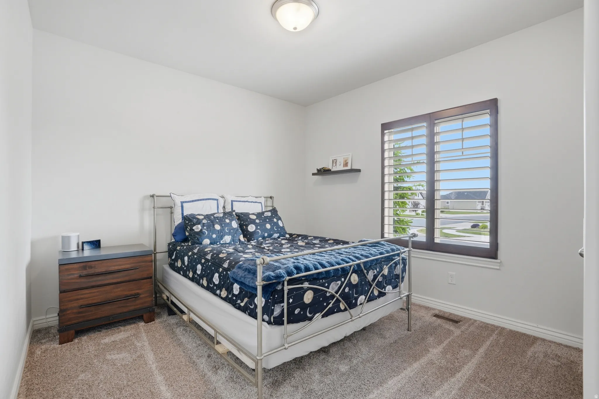 Bright bedroom featuring a large window with plantation shutters, a dark wood-finish window frame, and neutral carpeting