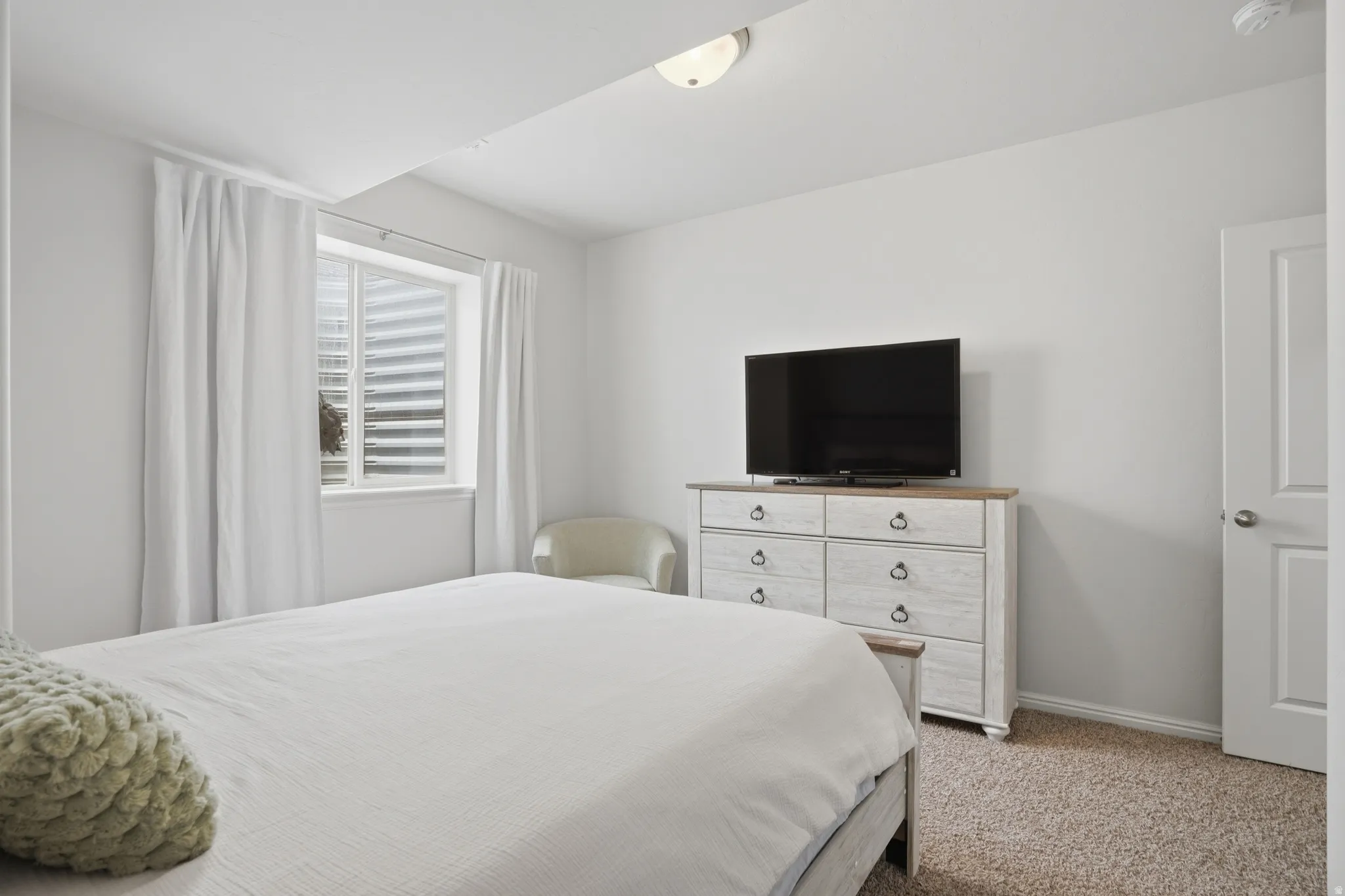Carpeted bedroom featuring a window with white blinds and curtains, white walls, and a ceiling light fixture