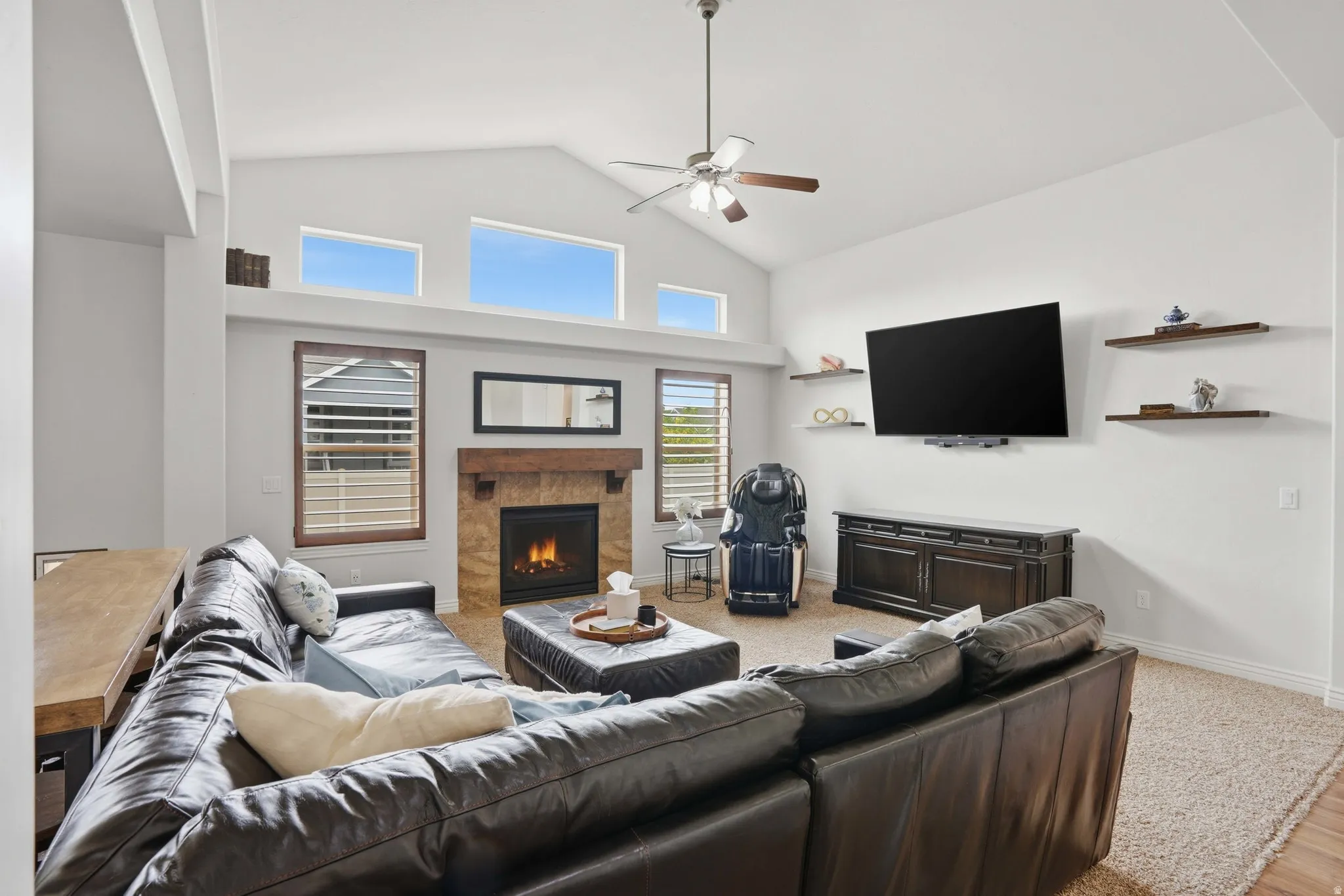 Vaulted ceiling living area featuring a stone-clad fireplace with a wood mantel, transom windows, and a ceiling fan