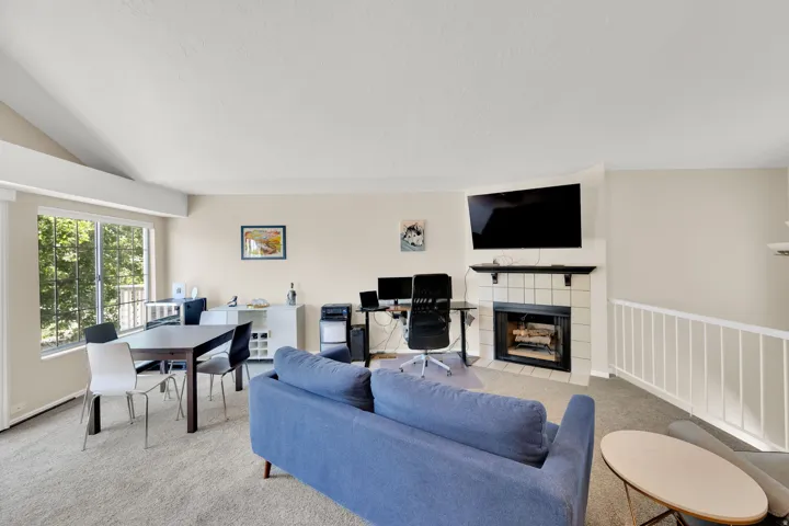 Spacious living area featuring a fireplace with a tiled surround, a large window, vaulted ceilings, and light-toned carpeting