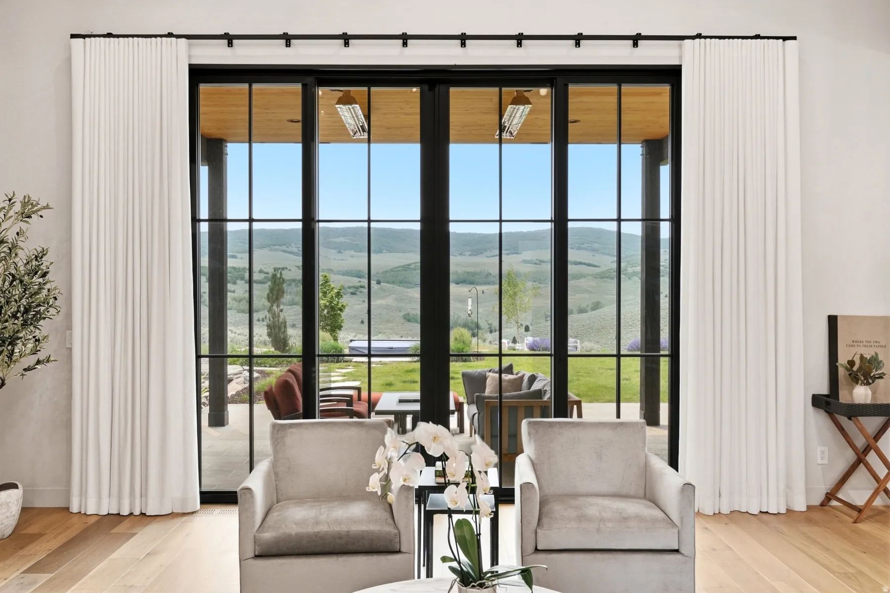 Expansive sliding glass doors with black grids open to an outdoor patio, framed by white drapery and light wood-finish flooring