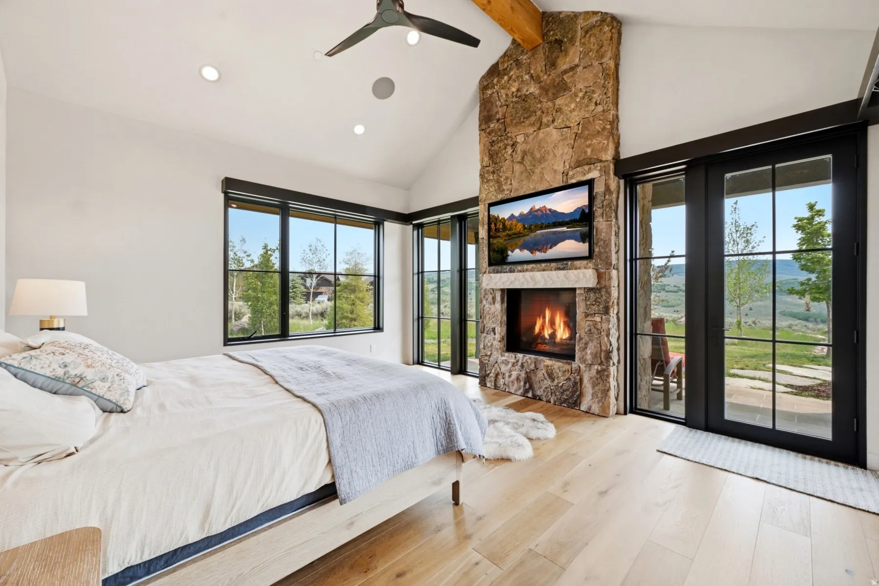 Vaulted ceiling room featuring a stone fireplace with a built-in television, light wood-finish flooring, and black-framed windows and glass doors