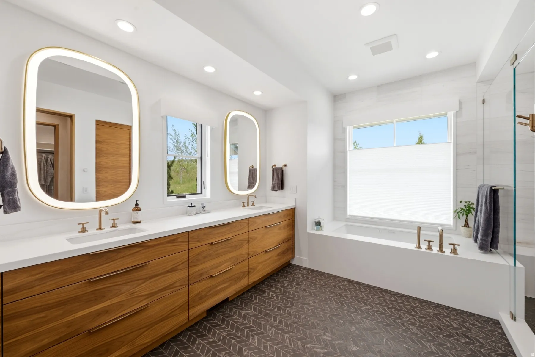 Herringbone tile flooring with a dual vanity featuring wood cabinetry, white countertops, and illuminated mirrors
