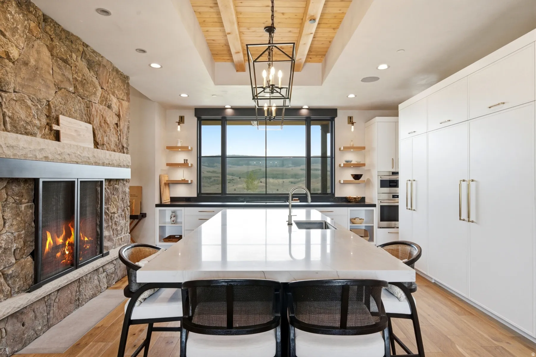 Spacious kitchen featuring a large central island with integrated sink, stone fireplace, extensive built-in white cabinetry, wood-finish flooring, and a beamed ceiling detail