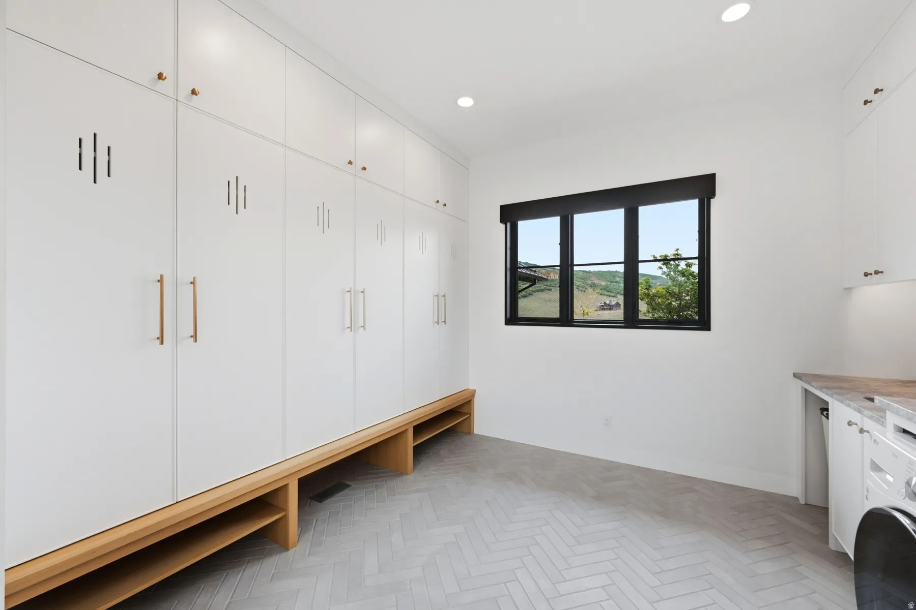 Mudroom featuring extensive white built-in cabinetry with gold-tone hardware, wood-finish bench seating, and herringbone pattern tile flooring