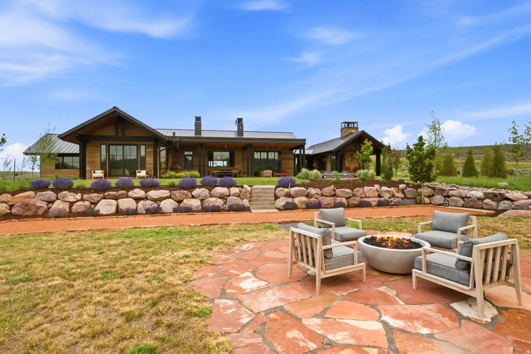 Expansive outdoor living area featuring a flagstone patio with a built-in fire pit, a gravel pathway, and a tiered rock retaining wall with planted shrubs