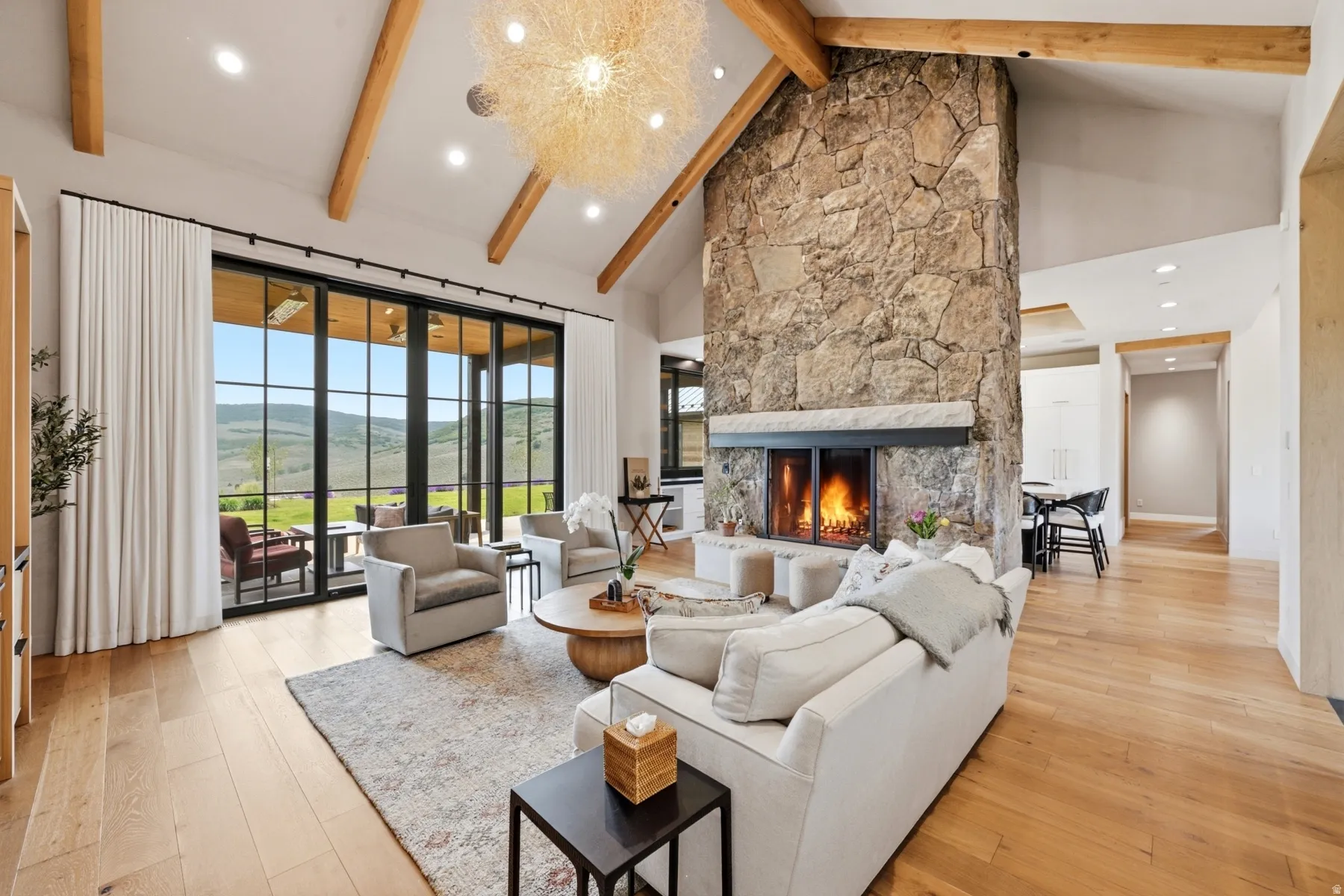 Spacious living area featuring a floor-to-ceiling stone fireplace, vaulted ceilings with exposed wood beams, and large glass sliding doors opening to an outdoor area