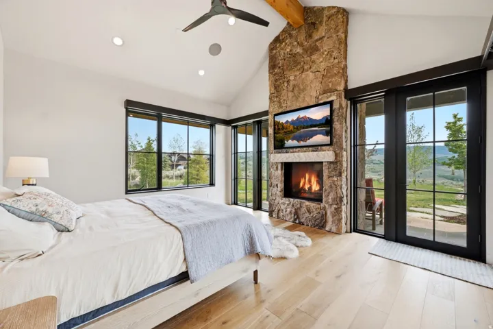 Vaulted ceiling room featuring a stone fireplace with a built-in television, light wood-finish flooring, and black-framed windows and glass doors