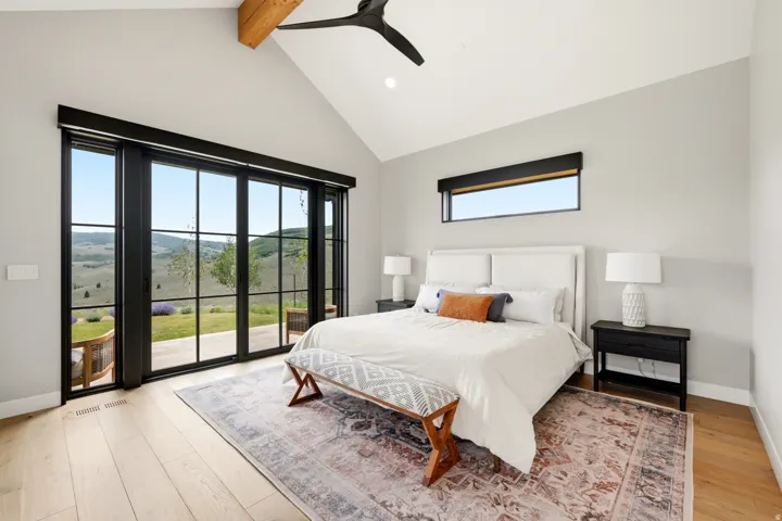 Spacious room featuring vaulted ceilings with exposed wood beams, expansive floor-to-ceiling black-framed sliding glass doors, light wood-finish flooring, and a horizontal transom window