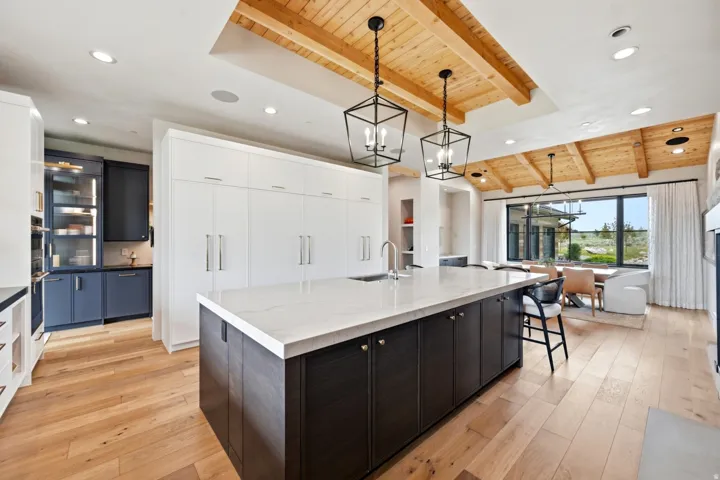 Spacious kitchen featuring a large island with a light-toned countertop, wood-finish flooring, white upper cabinetry, and a wood-paneled ceiling with exposed beams