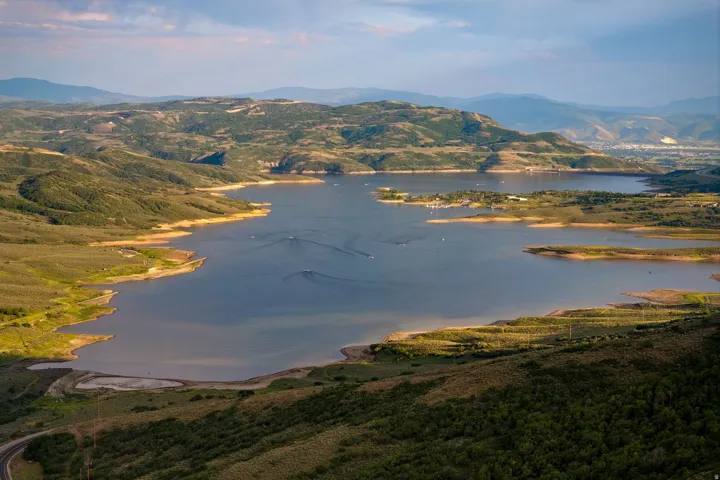 Expansive water body surrounded by green and brown hillsides, featuring distant mountains, a distant urban area, and multiple small boats on the water