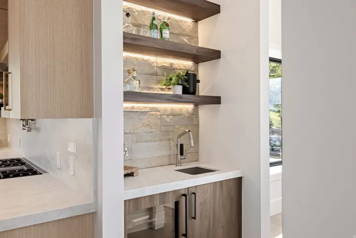 Wet bar featuring a stainless steel sink with a modern faucet, light stone accent wall, under-shelf lighting, floating wood shelves, and light wood-finish cabinetry with black hardware