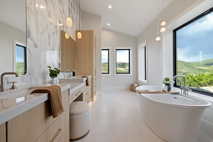 Spacious bathroom featuring a freestanding soaking tub, dual vanity with light wood-finish cabinetry, a large window showcasing natural scenery, and a marble-patterned accent wall