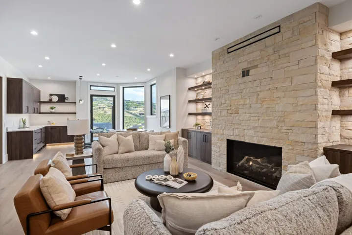 Living area featuring a stone-clad fireplace with an integrated linear vent, built-in shelving with accent lighting, wood-finish flooring, and a kitchen area with dark cabinetry and light countertops