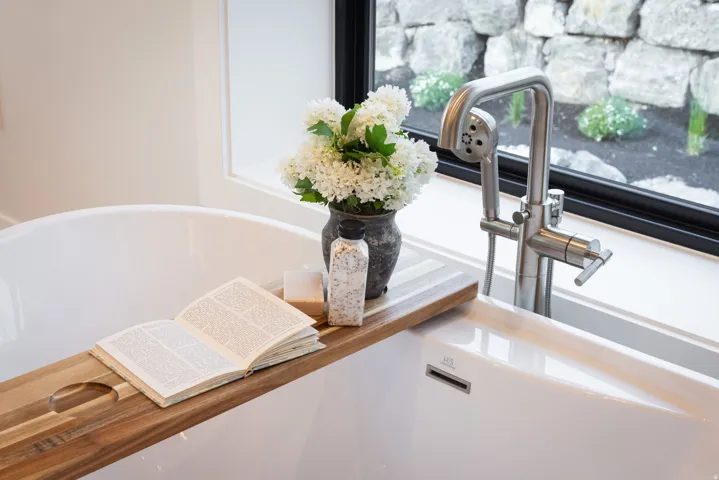 Freestanding soaking tub featuring a brushed nickel tub filler with hand shower, a large window with black trim, and a rock garden landscaping element visible outside