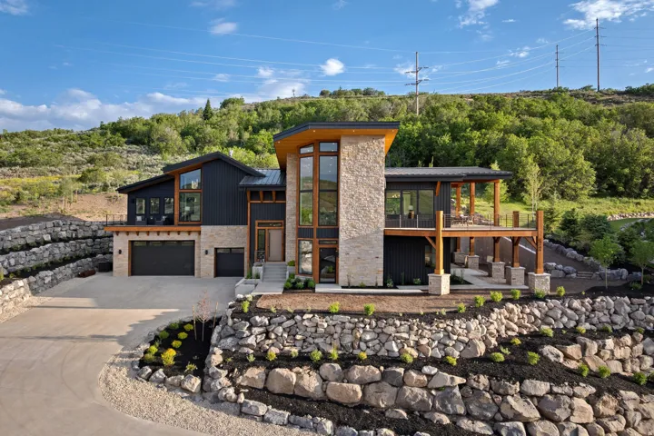 Modern architectural exterior featuring stone and dark vertical siding, multiple garages, and a prominent elevated deck with wood supports