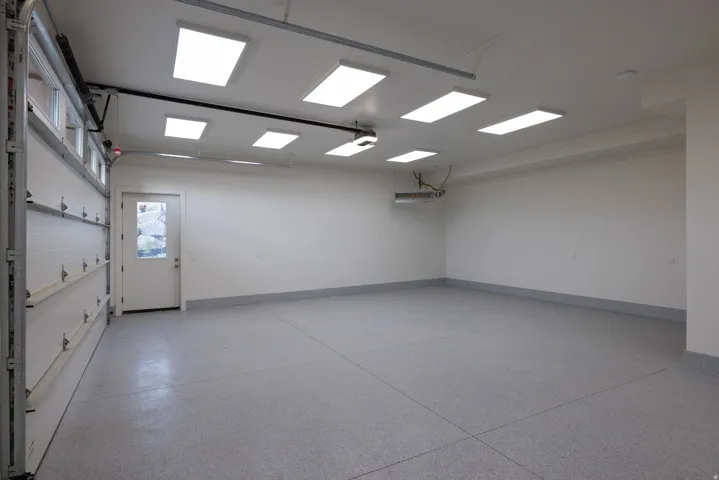 Spacious garage featuring a durable epoxy floor coating, multiple overhead fluorescent light fixtures, an insulated garage door with window inserts, a pedestrian door with a frosted glass panel, and a wall-mounted heating unit
