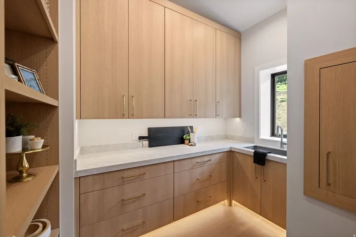 Custom built-in pantry featuring extensive wood-finish cabinetry, under-cabinet lighting, a built-in sink with a gooseneck faucet, light-toned countertops, and open shelving