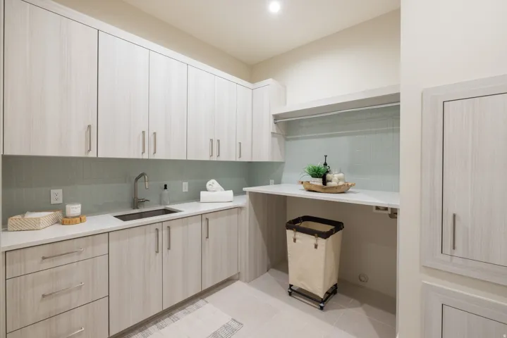 Laundry room featuring extensive light wood-finish cabinetry, a single basin sink with a modern faucet, quartz countertops, a pale blue tile backsplash, and built-in shelving with a hanging rod